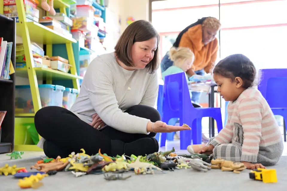 A teacher sitting on the floor playing with a young child and toy animals