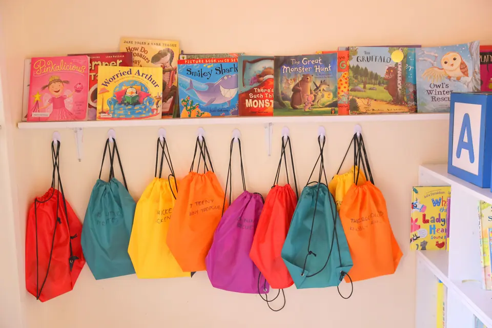 School bags hanging in a row beneath a shelf of books