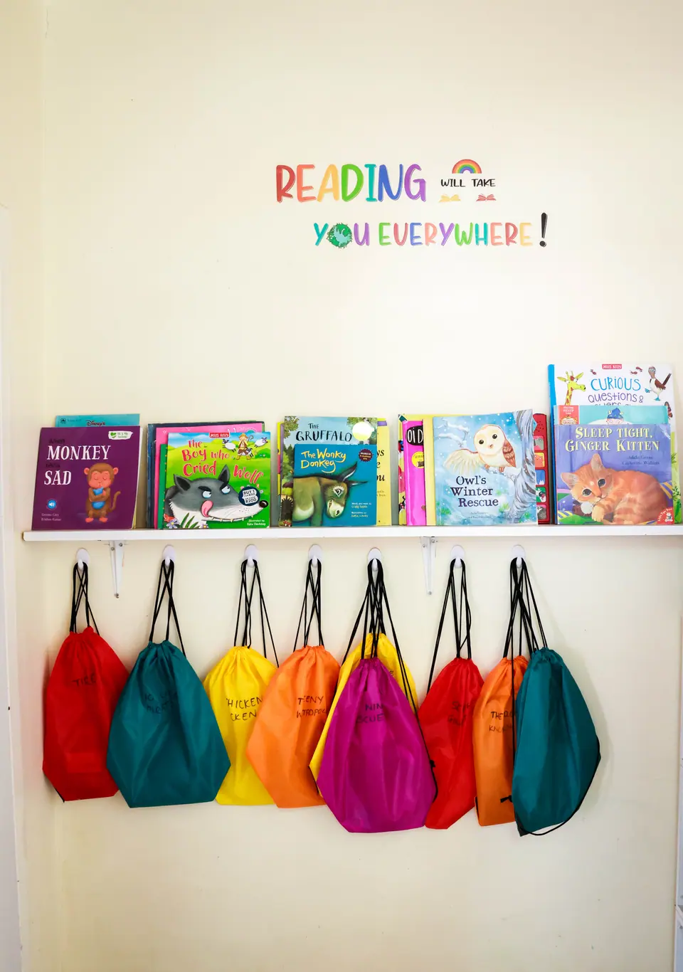 Colourful school bags hanging beneath a book shelf and a “Reading will take you everywhere” sign