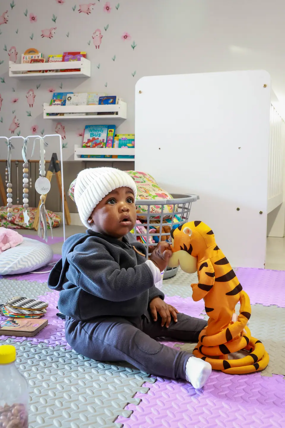 A baby in a knitted beanie sitting on soft play mats with a toy tiger