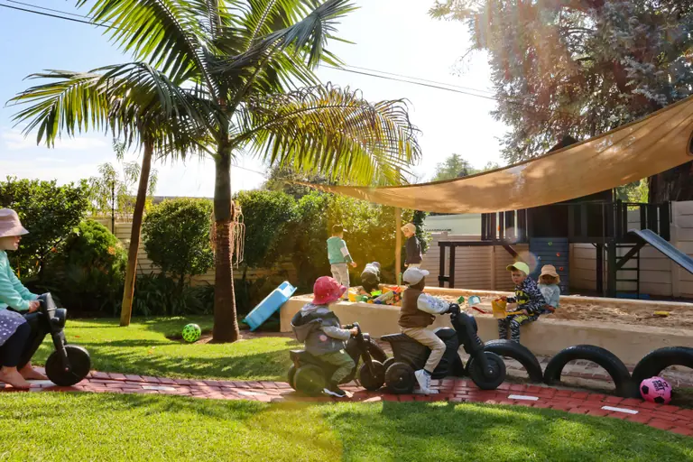 Children riding toy bikes along the garden path beneath a palm tree and shade sail